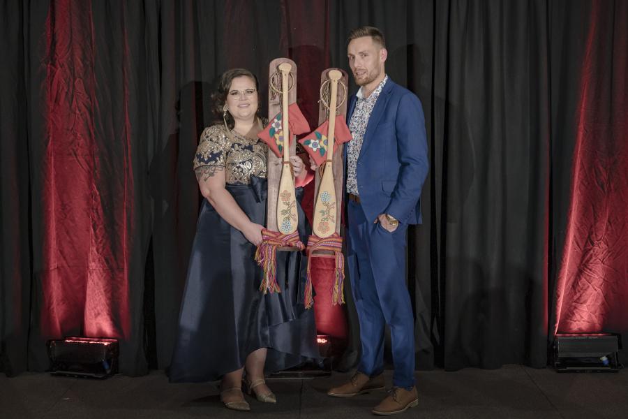 Janelle Desrosiers, wearing a blue dress, and Alex Hupe, wearing a blue suit, standing side by side holding their award paddles.